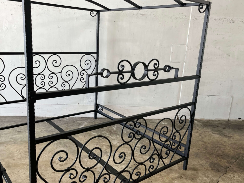 The Scroll Bed with Stockade by Metalbound: an ornate metal bed frame featuring intricate scrollwork and an integrated stockade, offering multiple attachment points for secure and versatile bondage play.